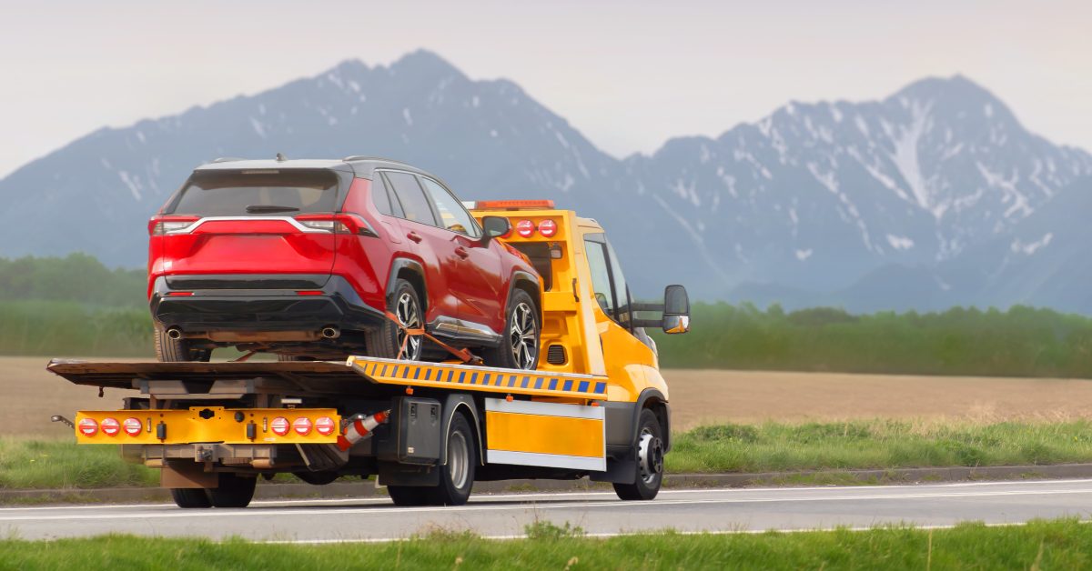 A red car being towed on a truck with mountains in the background